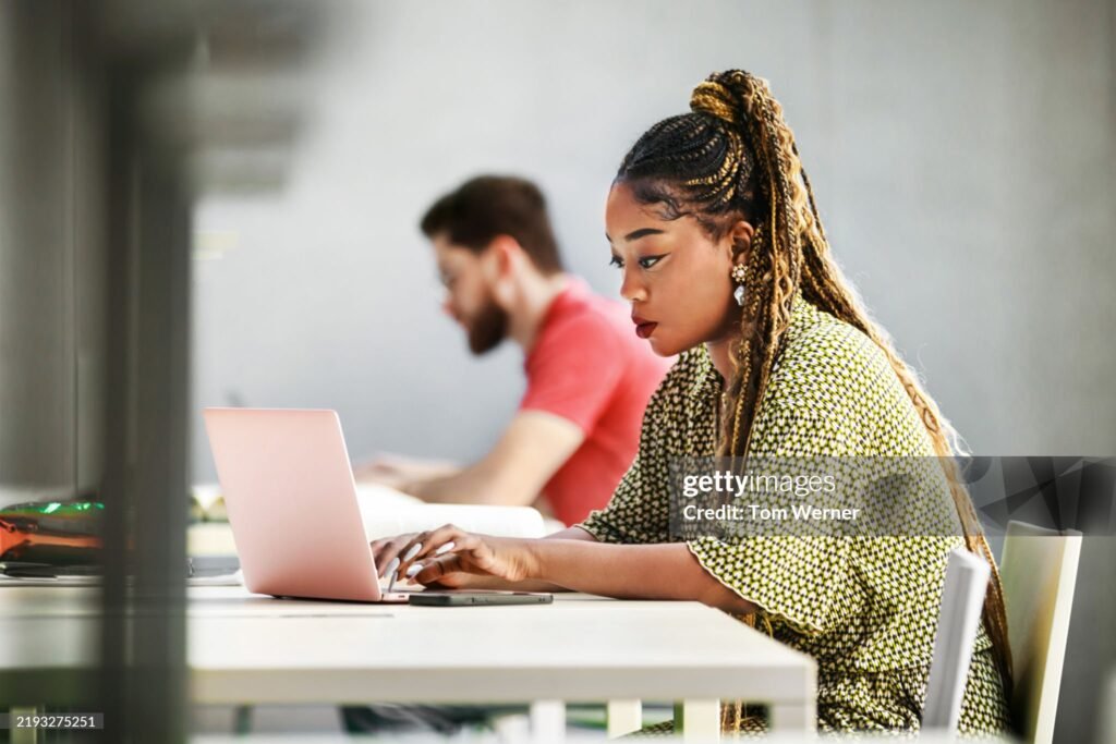 A young female student concentrates intently on her laptop while seated at a library. A blurred background creates a collaborative and productive atmosphere.